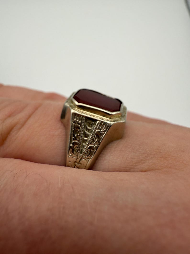 May include: A silver ring with a rectangular, deep red gemstone. The ring has an ornate, engraved design on the band. The gemstone is set in a gold-colored bezel. The ring is shown on a person's finger.