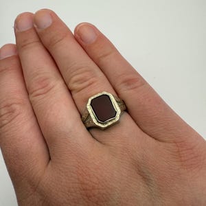 May include: A gold-toned signet ring with a rectangular, dark red stone. The ring has an ornate, engraved design around the stone and on the band. The ring is shown on a hand.