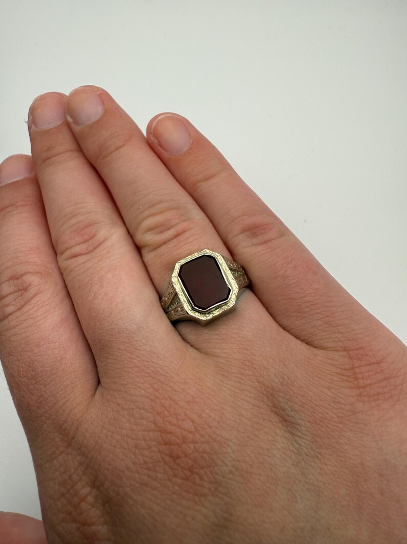 May include: A gold-toned signet ring with a rectangular, dark red stone. The ring has an ornate, engraved design around the stone and on the band. The ring is shown on a hand.
