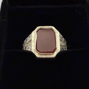 May include: A gold and silver ring with a rectangular, reddish-brown stone. The ring has an ornate design around the stone and on the band. The ring is set against a black background.