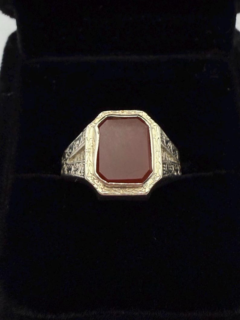 May include: A gold and silver ring with a rectangular, reddish-brown stone. The ring has an ornate design around the stone and on the band. The ring is set against a black background.
