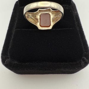 May include: A silver ring with a rectangular, reddish-brown stone set in a decorative bezel. The ring is displayed in a black velvet ring box, showcasing the jewelry's design and color contrast. The ring's band has a simple, polished finish.