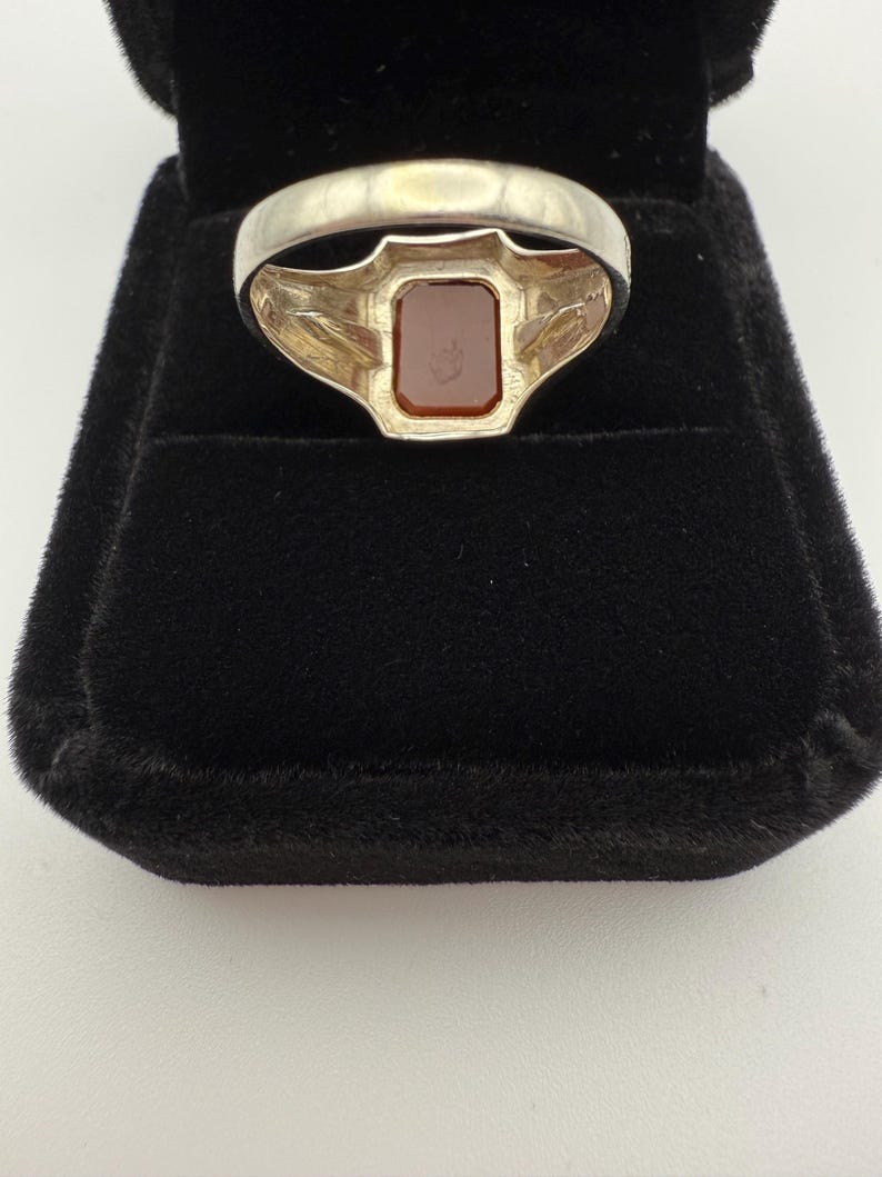 May include: A silver ring with a rectangular, reddish-brown stone set in a decorative bezel. The ring is displayed in a black velvet ring box, showcasing the jewelry's design and color contrast. The ring's band has a simple, polished finish.