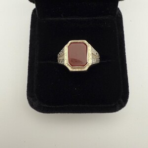 May include: A silver-toned ring with a rectangular, reddish-brown stone set in a gold-toned bezel. The ring has ornate detailing on the shoulders and is displayed in a black velvet ring box. Jewelry.