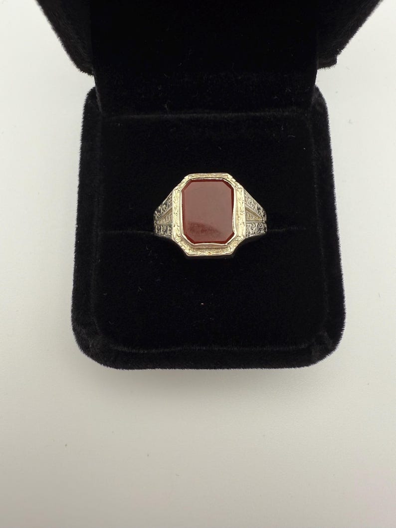 May include: A silver-toned ring with a rectangular, reddish-brown stone set in a gold-toned bezel. The ring has ornate detailing on the shoulders and is displayed in a black velvet ring box. Jewelry.