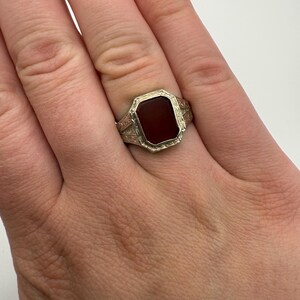 May include: A silver-toned ring with a rectangular, dark red stone. The ring has an ornate, engraved design around the stone and on the band. The ring is worn on a finger.