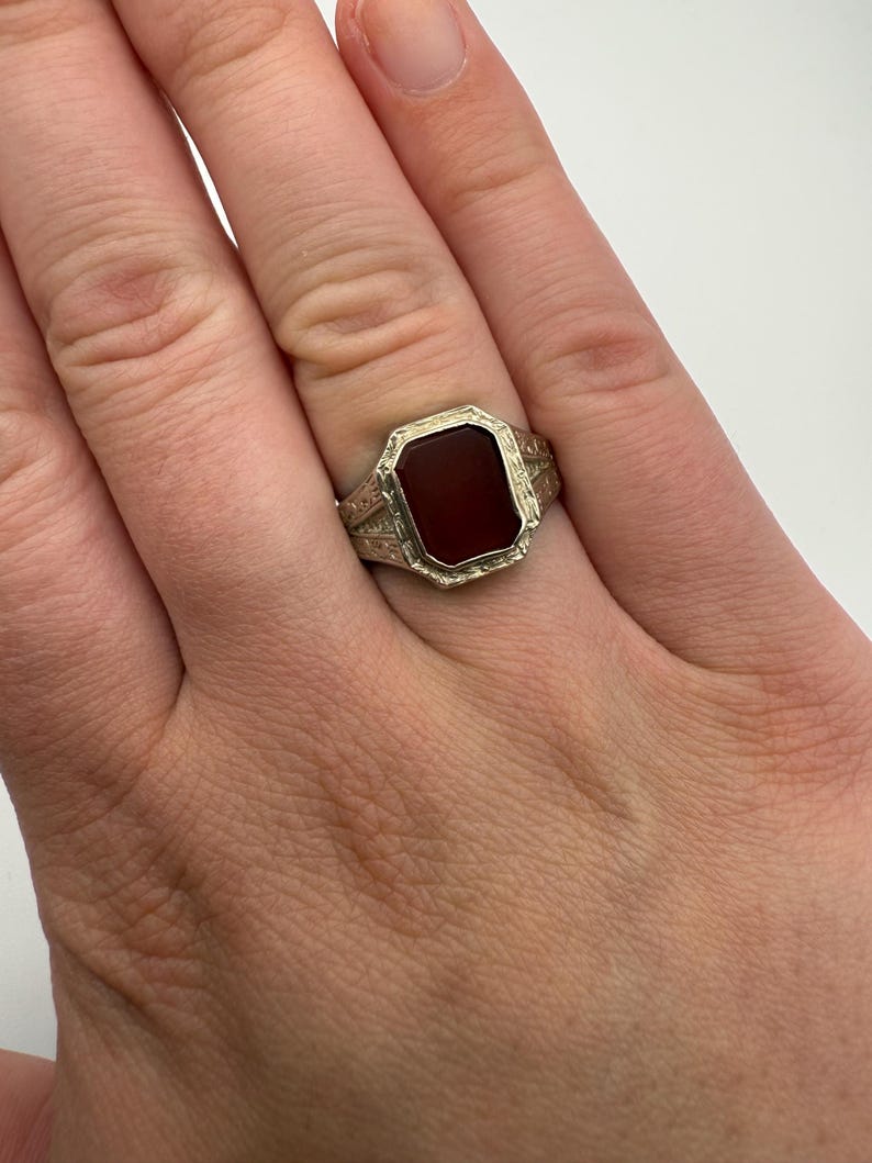 May include: A silver-toned ring with a rectangular, dark red stone. The ring has an ornate, engraved design around the stone and on the band. The ring is worn on a finger.