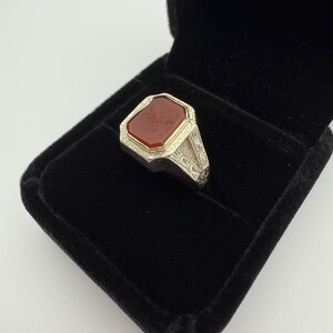 May include: A silver ring with a rectangular, reddish-brown stone set in a decorative bezel. The ring is displayed in a black velvet ring box, showcasing the intricate details of the metalwork and the stone's color.
