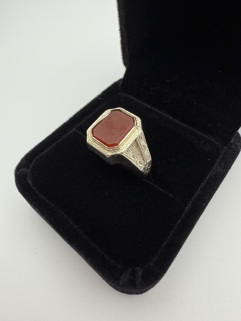 May include: A silver ring with a rectangular, reddish-brown stone set in a decorative bezel. The ring is displayed in a black velvet ring box, showcasing the intricate details of the metalwork and the stone's color.