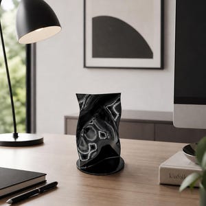 May include: A glossy black vase with a unique, twisted design sits on a wooden desk. The vase has a wide base and a flared top. A black desk lamp and a framed abstract art piece are in the background.