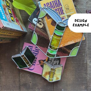 May include: Stained glass book-shaped ornaments in shades of green, yellow, and blue, with silver-colored metal edging. The ornaments are placed on top of vintage Agatha Christie novels. The text "DESIGN EXAMPLE" is visible.