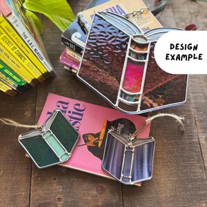 May include: Stained glass book-shaped ornaments in shades of purple, green, and brown, with silver-colored metal edging. The ornaments are displayed near a stack of books, including Agatha Christie novels. The text "DESIGN EXAMPLE" is visible.