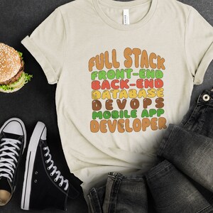 May include: A beige t-shirt with the text "FULL STACK FRONT-END BACK-END DATABASE DEVOPS MOBILE APP DEVELOPER" in colorful, rounded letters.