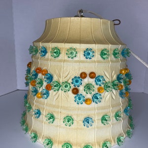 Może przedstawiać: Kremowy, zabytkowy klosz lampy z teksturowaną powierzchnią i kwiatowymi akcentami. Klosz jest ozdobiony niebieskimi, zielonymi i pomarańczowymi plastikowymi ozdobami w kształcie kwiatów. Do góry przymocowany jest biały przewód i metalowy haczyk.