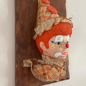 May include: A wall-mounted clown head plaque featuring a textured orange wig, a plaid cone hat with orange pom-poms, and a ruffled collar. The clown has painted facial features including blue and yellow diamond shapes, a red nose, and white lips.