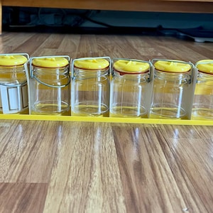 May include: Six small, clear glass jars with yellow lids and metal handles are arranged on a yellow tray. The jars are empty, and the tray sits on a wooden surface. The jars are suitable for storing spices or small items.