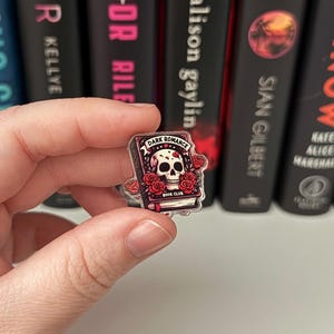 Può includere: Uno spillo in acrilico trasparente con un design di club di lettura di romance oscuro. La copertina del libro presenta uno scheletro con rose rosse e il testo "Dark Romance Book Club".