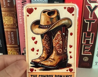 May include: A rectangular sticker featuring a cowboy hat and boots, with red heart accents. The sticker has the text "THE COWBOY ROMANCE" at the bottom. The hat and boots are brown with decorative details.