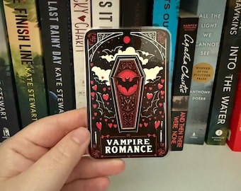 May include: A rectangular sticker with a black background and a coffin design. The coffin is red with a bat in the center. The sticker has the words "VAMPIRE ROMANCE" at the bottom, with hearts and bats as decorative elements.