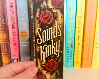 Könnte beinhalten: Ein schwarzes Lesezeichen mit goldenem Hintergrund und roten Rosen. Der Text "Sounds Kinky" ist in einer stilisierten Schrift geschrieben.
