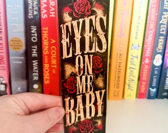 Könnte beinhalten: Ein schwarzer Lesezeichen mit dem Text "EYES ON ME BABY" in einer Vintage-Schriftart, verziert mit roten Rosenillustrationen. Das Lesezeichen wird vor einem Regal mit bunten Büchern gehalten.
