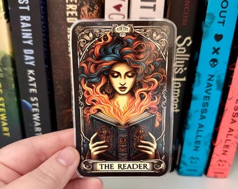 May include: A rectangular sticker featuring a woman with fiery hair and a book, labeled "THE READER." The artwork has a vintage tarot card aesthetic, with ornate borders and a dark color palette. The sticker is held in front of a bookshelf.