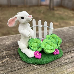 May include: A white ceramic bunny figurine standing in a miniature garden setting. The bunny is surrounded by green leafy plants and pink flowers. A white picket fence is behind the bunny.