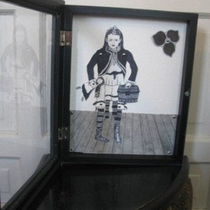 May include: A black framed shadow box with a black and white illustration of a girl holding a doll and a suitcase. The girl is wearing a black jacket and a white dress. The background is a light gray.