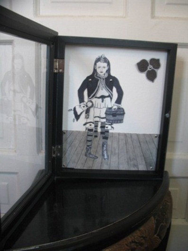 May include: A black framed shadow box with a black and white illustration of a girl holding a doll and a suitcase. The girl is wearing a black jacket and a white dress. The background is a light gray.