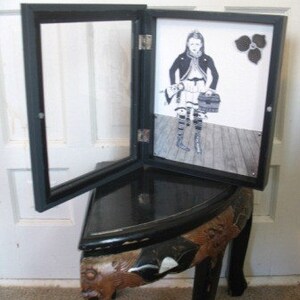 May include: A black wooden picture frame with a glass front and a hinged door. The frame is open to reveal a black and white photograph of a girl wearing a dress and holding a purse. The photo is on a wooden surface.