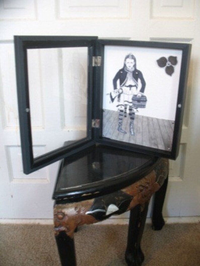 May include: A black wooden picture frame with a glass front and a hinged door. The frame is open to reveal a black and white photograph of a girl wearing a dress and holding a purse. The photo is on a wooden surface.
