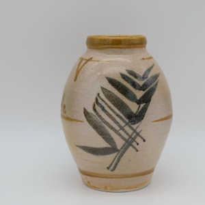 May include: A beige ceramic vase with a rounded body and a narrow neck. The vase features a hand-painted design of stylized dark green and black leaves and lines. The rim and base have a brown band.