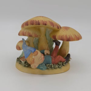 Könnte beinhalten: Keramikfigur eines Zwergs, der unter einer Gruppe von Pilzen ruht. Der Zwerg trägt einen blauen Hut, ein grünes Hemd und eine rote Hose. Die Pilze sind beige mit rotbraunen Kappen.