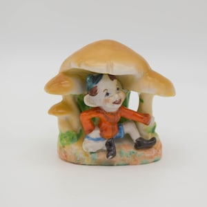 Peut inclure: Figurine en céramique d'un elfe assis sous un champignon. L'elfe porte une chemise orange, un short bleu et un bonnet bleu. Le champignon a un chapeau beige et une tige blanche. La base a des accents verts et bruns.
