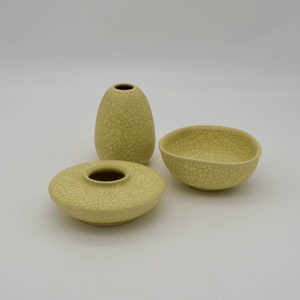 May include: Three ceramic vessels in a pale yellow hue with a crackle glaze. The set includes a small vase, a shallow bowl, and a low, round dish. The objects are arranged on a white background.