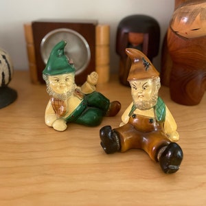 Może przedstawiać: Dwie ceramiczne figurki krasnali ogrodowych. Jeden krasnal ma na sobie zielony kapelusz i ogrodniczki, drugi brązowy kapelusz i ogrodniczki. Obaj mają białe brody i siedzą. Krasnale stoją na drewnianej półce.