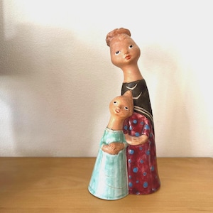 May include: A ceramic figurine depicting two figures, one taller and one shorter, standing close together. The taller figure wears a black dress with white stripes and the shorter figure wears a light blue dress. The taller figure's dress has red polka dots.