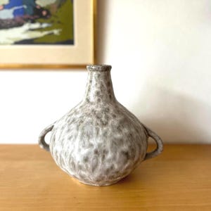 May include: A grey ceramic vase with a textured surface and two small handles. The vase has a bulbous body and a narrow neck. The vase is on a wooden surface.