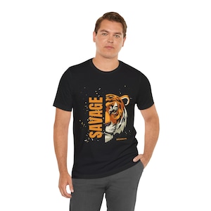 Puede incluir: Camiseta negra con la cara de un tigre a rayas naranjas y blancas. La palabra "SAVAGE" está impresa verticalmente en naranja, con "BROOKLYN" debajo. La camiseta está hecha de un material suave.