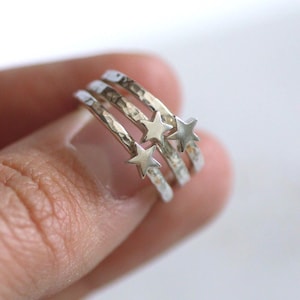 May include: Three silver rings with a hammered texture, each featuring a small star. The rings are stacked, creating a layered effect. The rings are held in a hand against a white background.