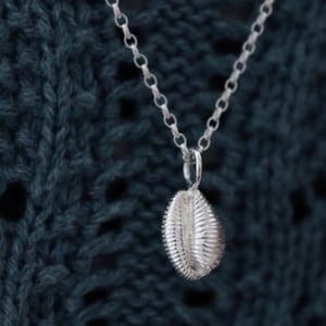 May include: A silver necklace featuring a seashell pendant. The pendant, with detailed ridges, hangs from a fine chain. The background is a dark teal knitted fabric.