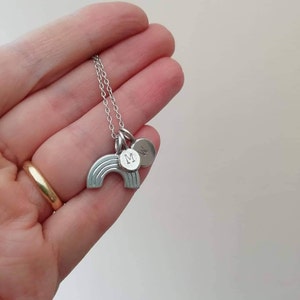 The rainbow charm is shown with two personalised pebble charms with initials. They are hanging on a chain draped over a hand. The rainbow is a little larger than the pebbles.