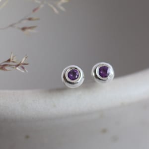 May include: A pair of silver stud earrings. Each earring has a small, round, deep purple gemstone set in a silver bezel. The earrings are displayed on a white surface. The earrings are a simple, elegant design.