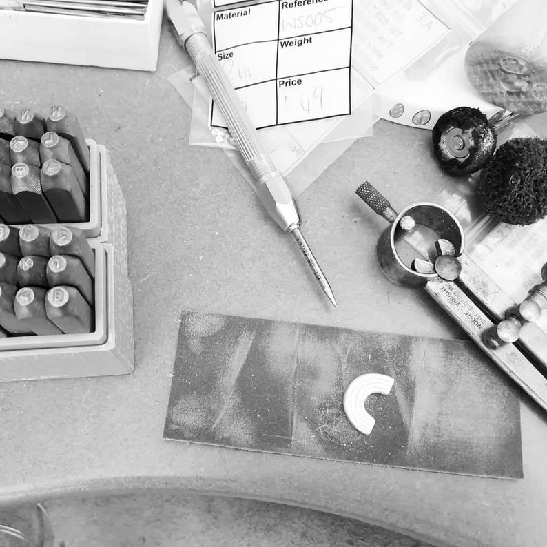 A rainbow charm is shown on a jewellery bench with the tools needed for its making. There are letter punches at the ready to add text to the pendant.