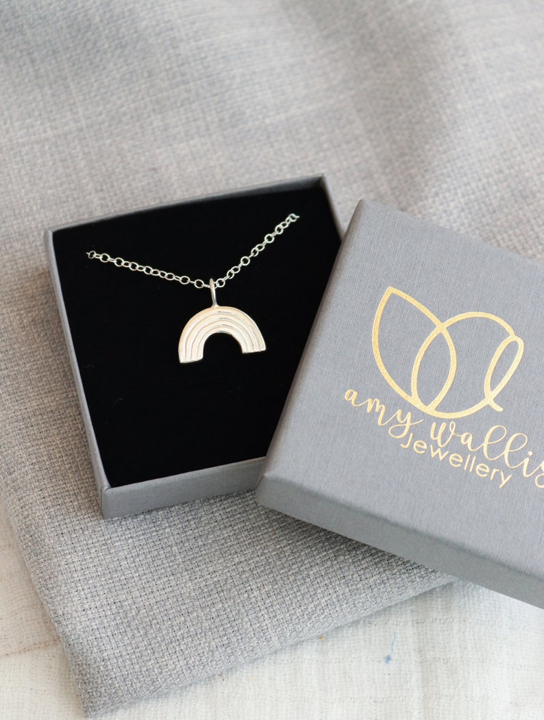 The silver rainbow charm is on a fine chain displayed inside a branded grey jewellery box.