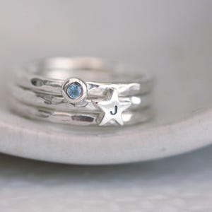 May include: A stack of three silver rings with a hammered texture. One ring features a small, round, light blue gemstone, while another ring is adorned with a silver star charm stamped with the letter J in a dark, clear font.