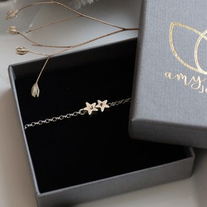A dainty silver bracelet is displayed in a grey branded jewellery box. The bracelet has two stars, you can choose the number and the initials that go on each star. All of this is on top of a white star dish.