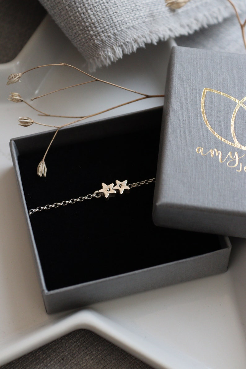 A dainty silver bracelet is displayed in a grey branded jewellery box. The bracelet has two stars, you can choose the number and the initials that go on each star. All of this is on top of a white star dish.