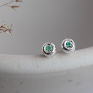 May include: A pair of small, round silver stud earrings with a bezel-set, vibrant green gemstone in the center of each. The earrings are displayed on a white, textured surface, with a blurred background.