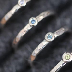 May include: Close-up of several silver rings with small, round, light blue gemstones. The rings have a hammered texture and a simple design. The gemstones are set in a bezel setting. The rings are arranged diagonally.